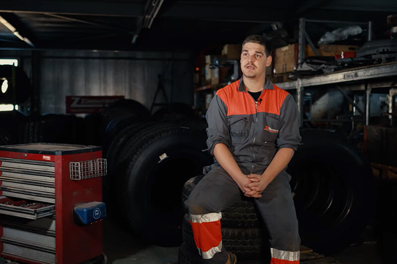 Vorschaubild Lehrlingsvideo mit Lehrling, der auf Reifenstapel sitzt. Darüber Play-Button