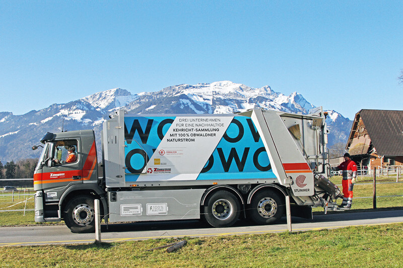 Ein Kehrrichtfahrzeug steht auf einer Landstraße vor einem Alpenpanorama, mit einem Arbeiter darauf.